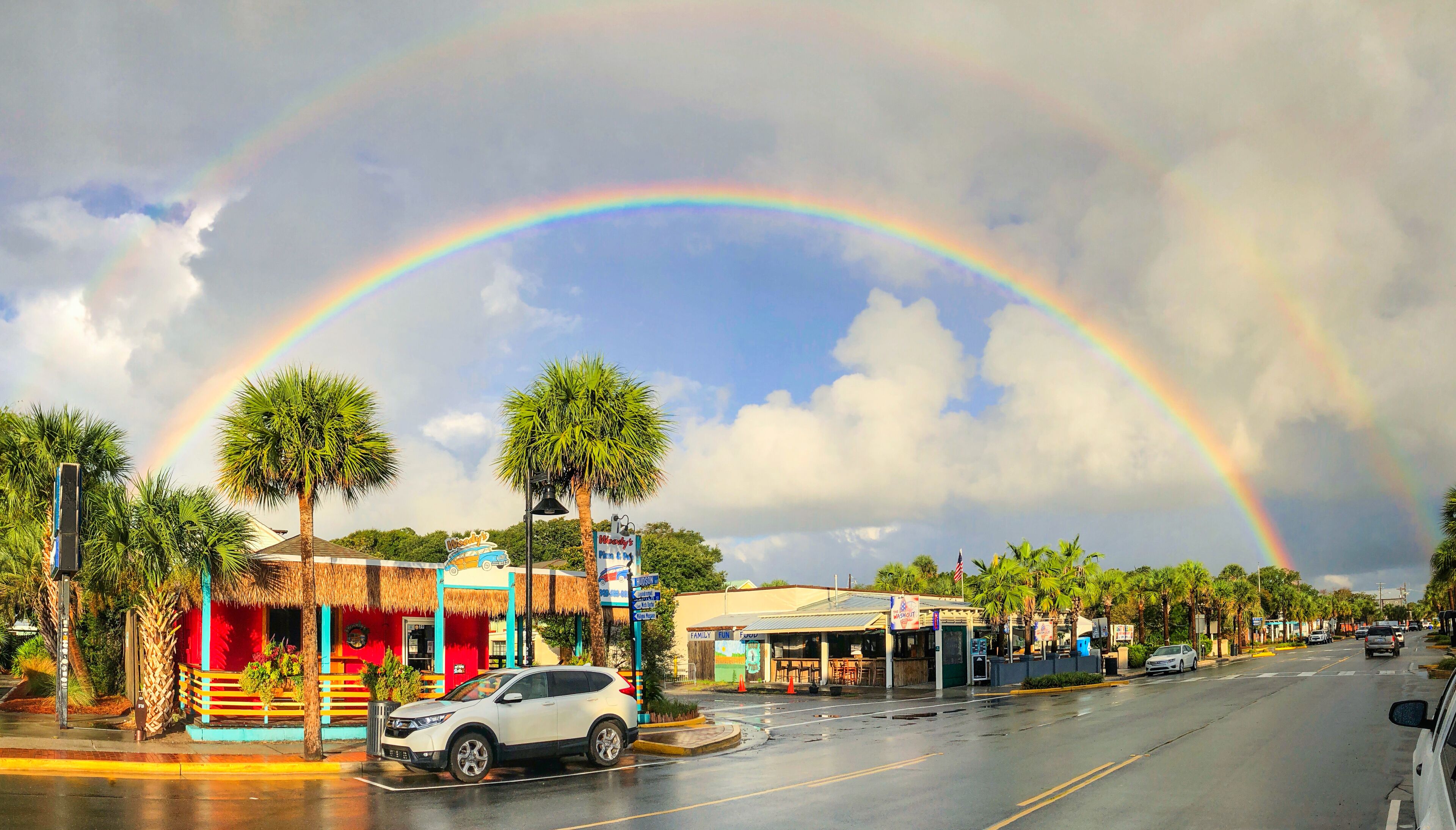 A street with a white SUV parked by the curb, palm trees lining the road, and a rainbow arching across the sky.