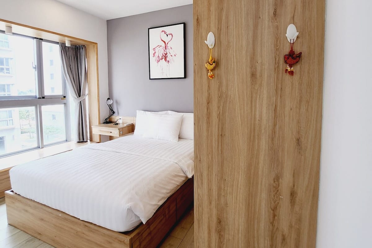 Een moderne slaapkamer met een houten bedframe, een nachtkastje, een bijzettafeltje, een kunstwerk in een lijst en een raam met gordijnen.