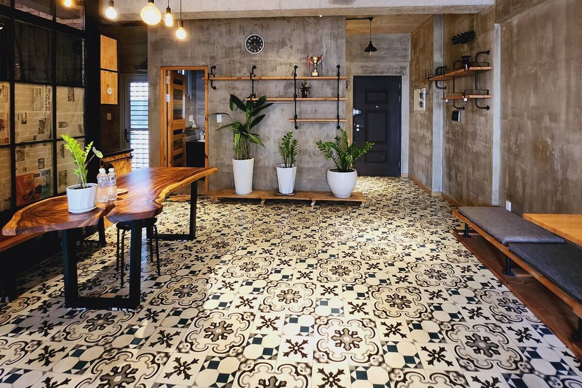 Een ruime kamer met een hoge plafond, zichtbaar leidingwerk en een vloer met patroon. Er staat een houten tafel met stoelen, planten in potten en een plafondventilator.