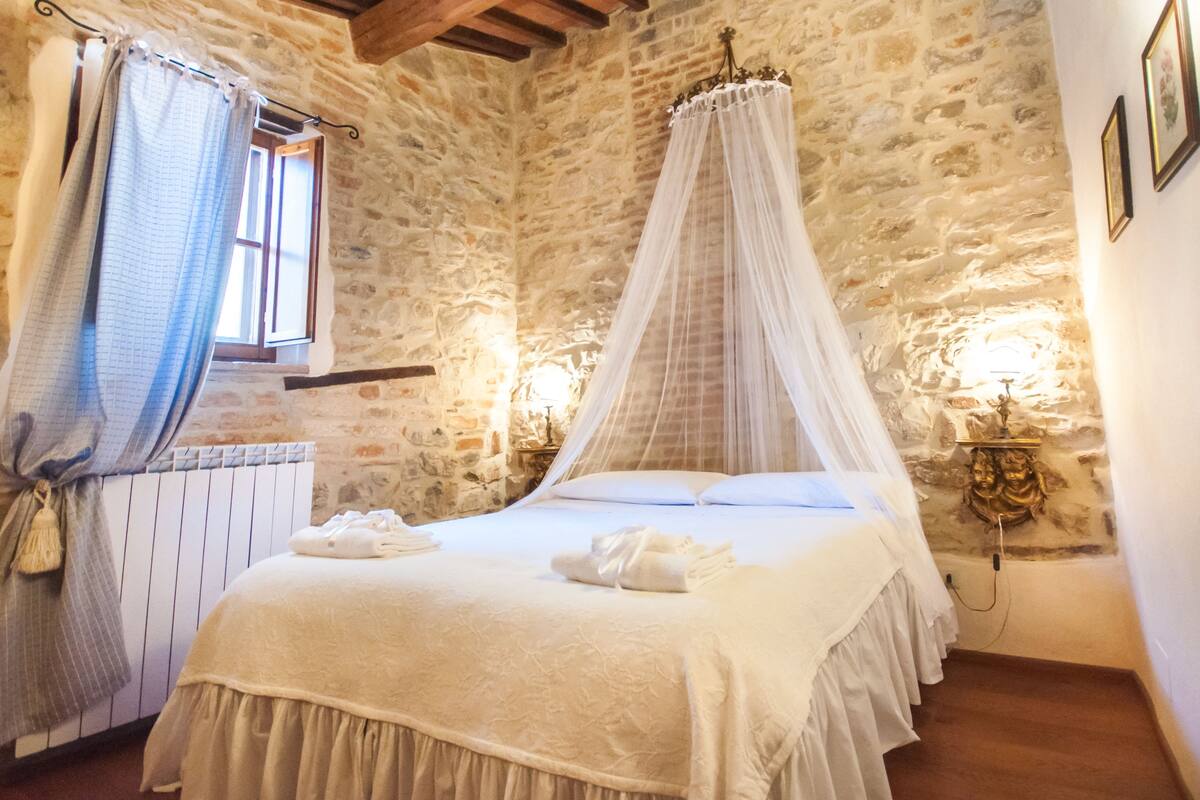 A bedroom with a stone wall, a four-poster bed, a window with curtains, and wall-mounted lights.