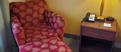 A hotel room with a red patterned armchair, a wooden nightstand with a lamp, a phone, and a digital clock.