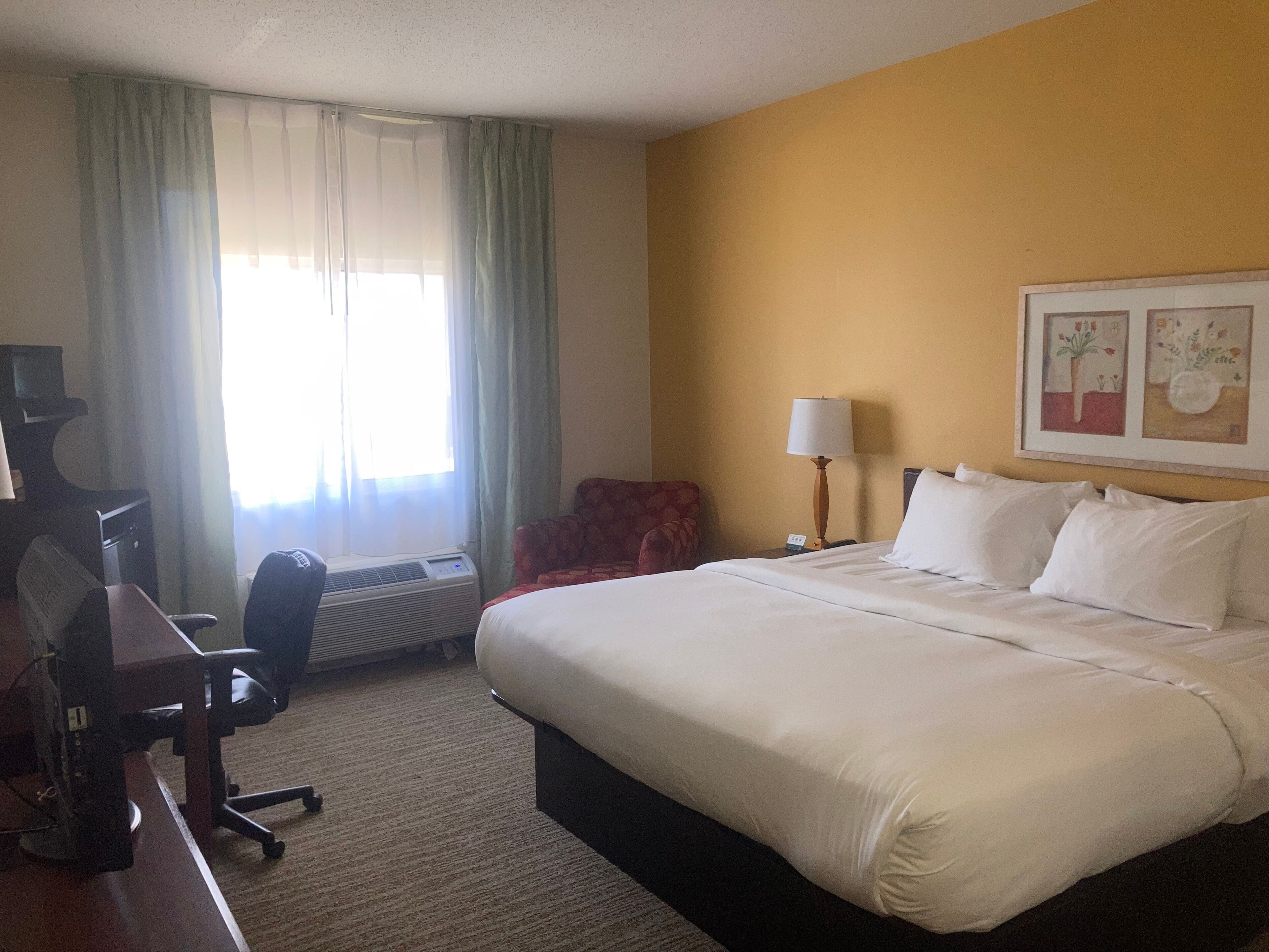 Ein Hotelzimmer mit einem Bett, einem Schreibtisch mit Computer, einem Sessel, einer Lampe und einem Fenster mit Vorhängen.