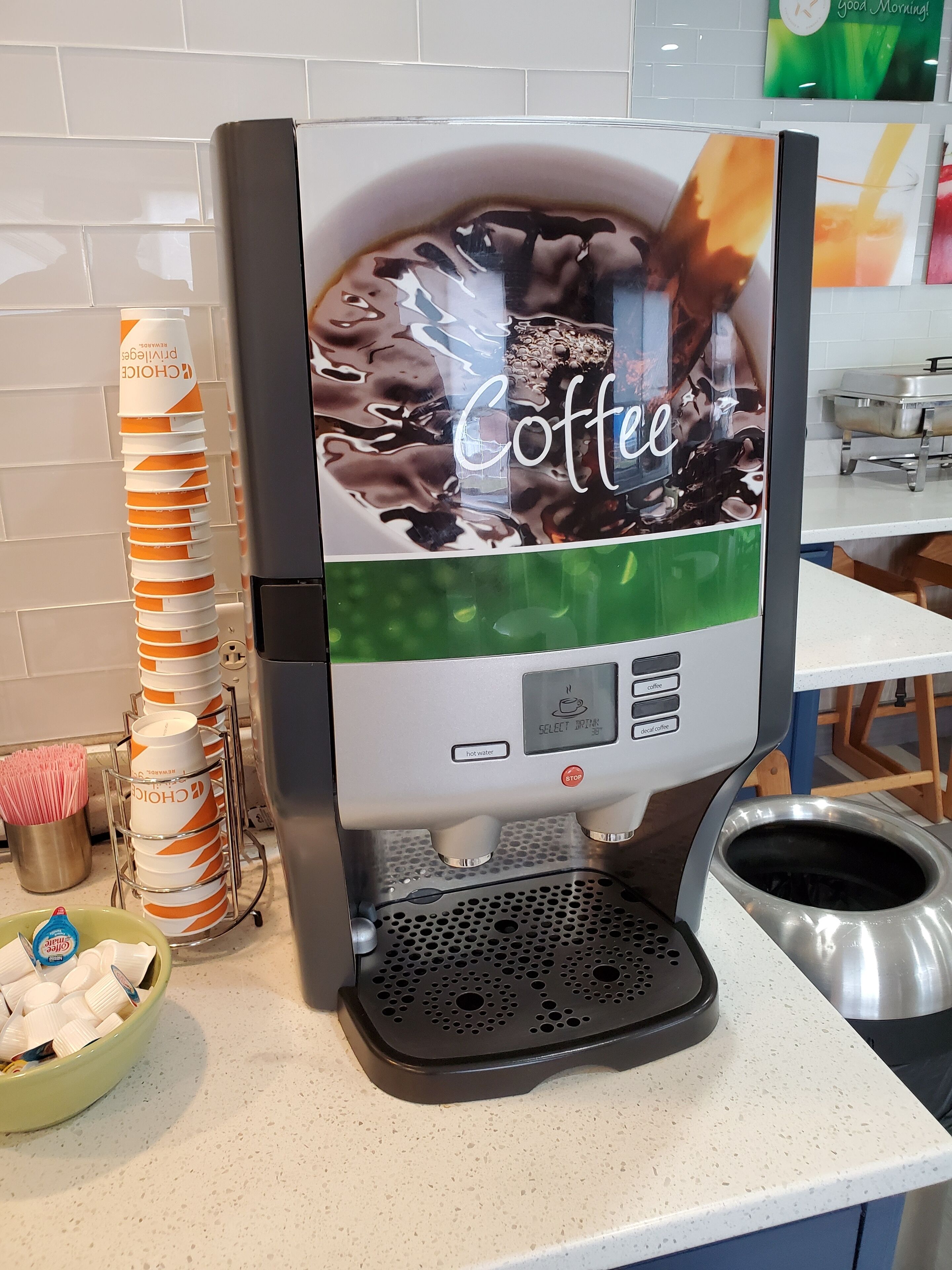 Una máquina de café con diseño temático de café, una pila de vasos desechables y un recipiente con cápsulas de café.