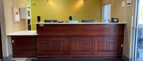 A reception area with a wooden counter, a yellow wall, and a black mat with social distancing markings.