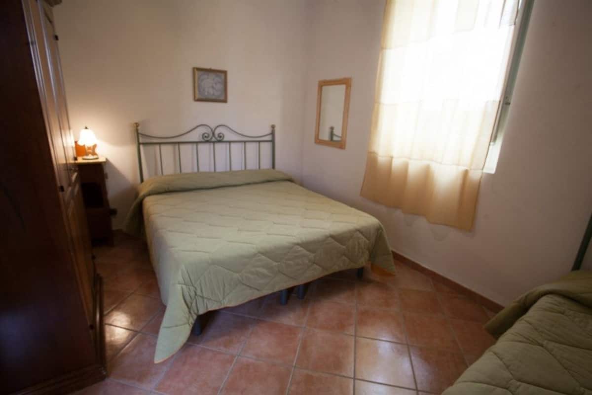 Een slaapkamer met een bed, nachtkastje, spiegel en raam met gordijnen.