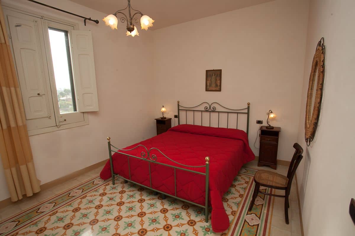 Een slaapkamer met een bed, een stoel, een nachtkastje, een lamp, een raam met gordijnen en een spiegel aan de muur.