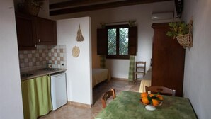 A compact kitchen with a dining area, featuring a small table with a fruit bowl, a window with curtains, and a kitchenette with a refrigerator. - Agriturismo Baglio Donna Santa (Paceco)
