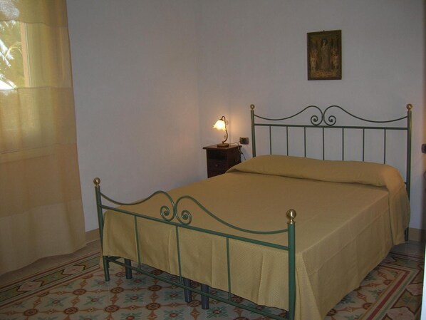 A single bed with a metal frame and a beige bedspread in a room with patterned floor tiles and a small bedside table with a lamp. - Agriturismo Baglio Donna Santa (Paceco)