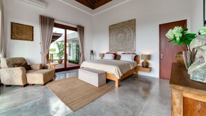 A spacious bedroom with a large bed, a sitting area with a chair, a small table, and a decorative wall piece. - Villa Daana Uluwatu (Ungasan)