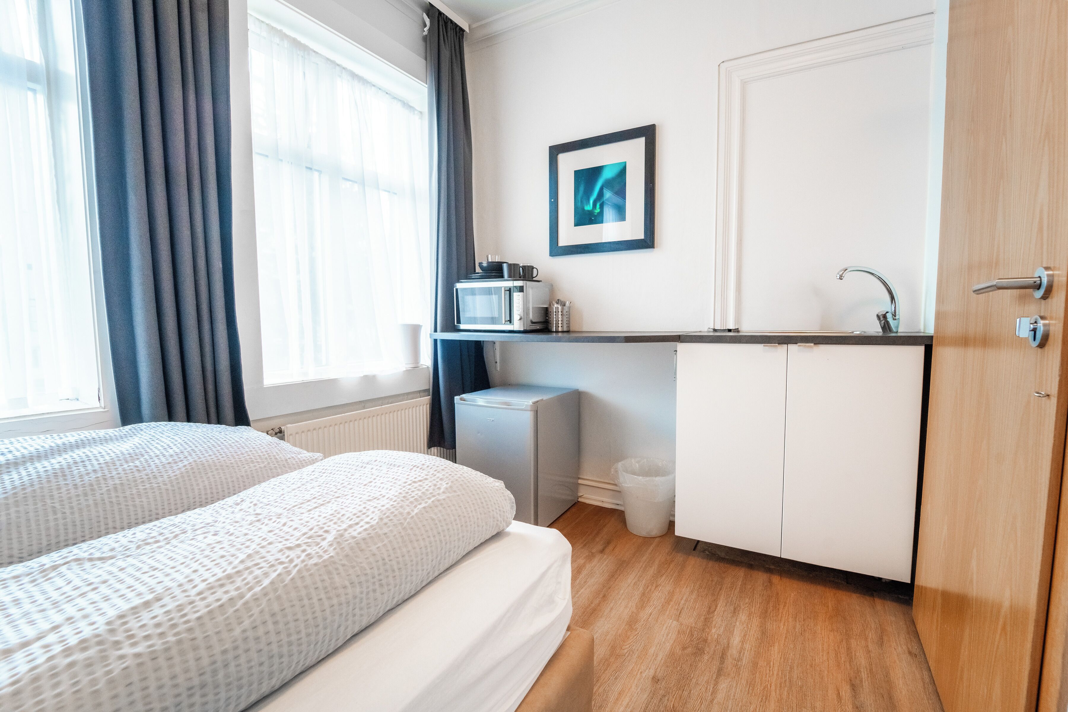 Een hotelkamer met een bed, een kleine kitchenette, een witte deur en een raam met gordijnen.