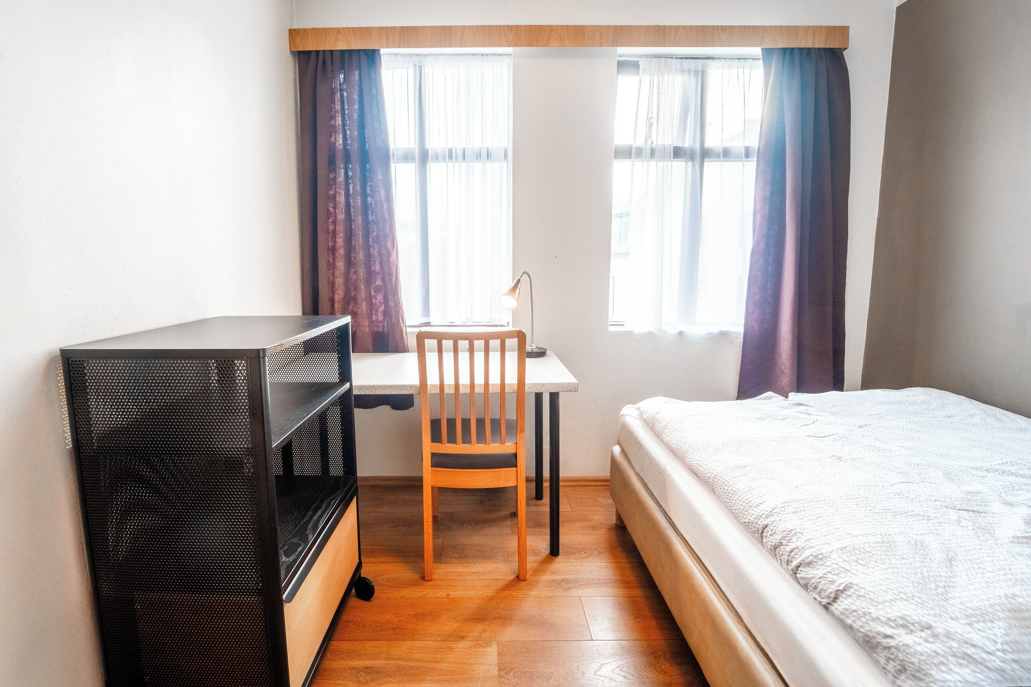 A compact room with a bed, a desk with a chair, and a black storage unit.