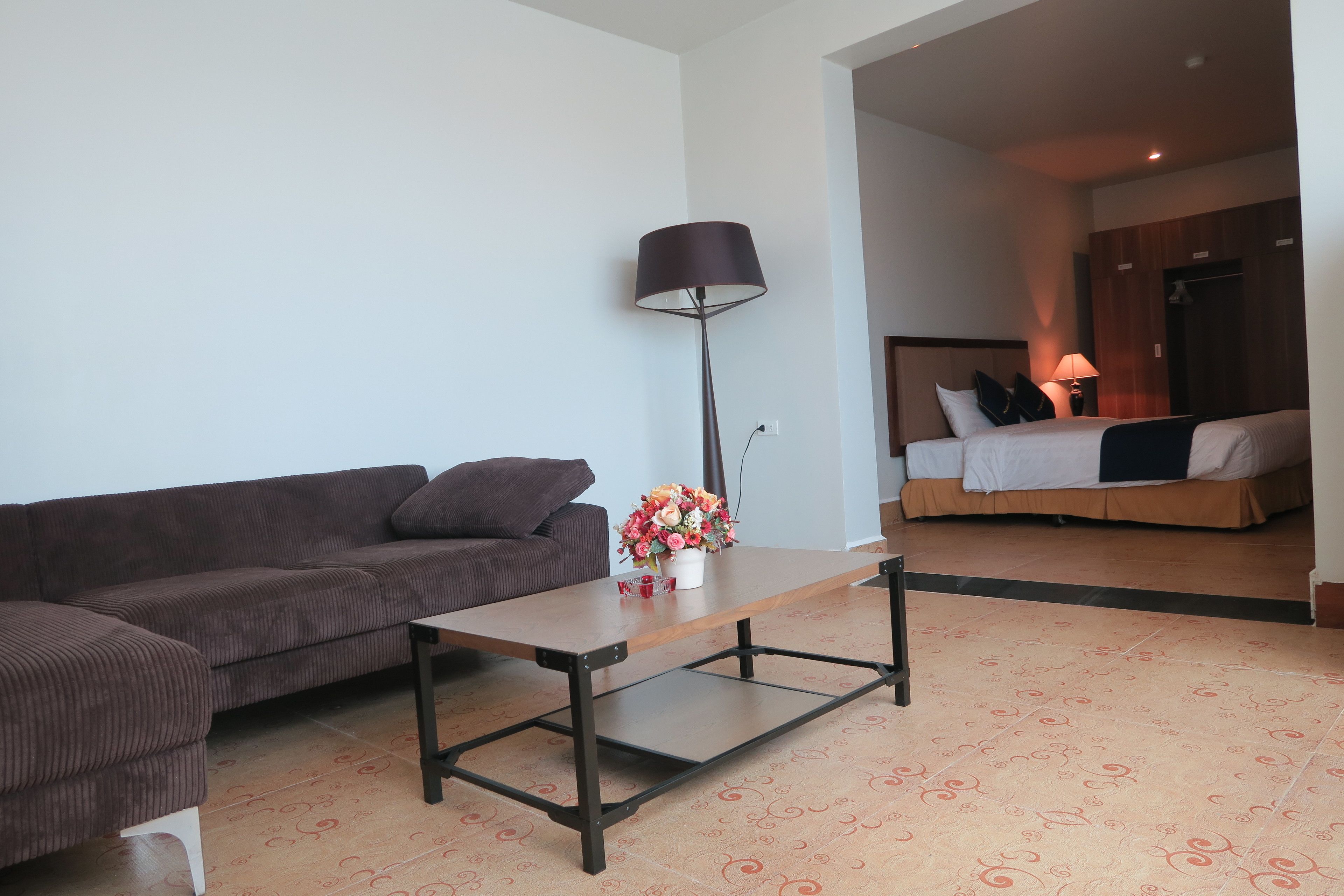 Un salon moderne avec un canapé, une table basse et une lampe. On aperçoit, au fond, un lit avec une tête de lit et des oreillers.