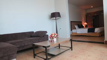Un salon moderne avec un canapé, une table basse et une lampe. On aperçoit, au fond, un lit avec une tête de lit et des oreillers.
