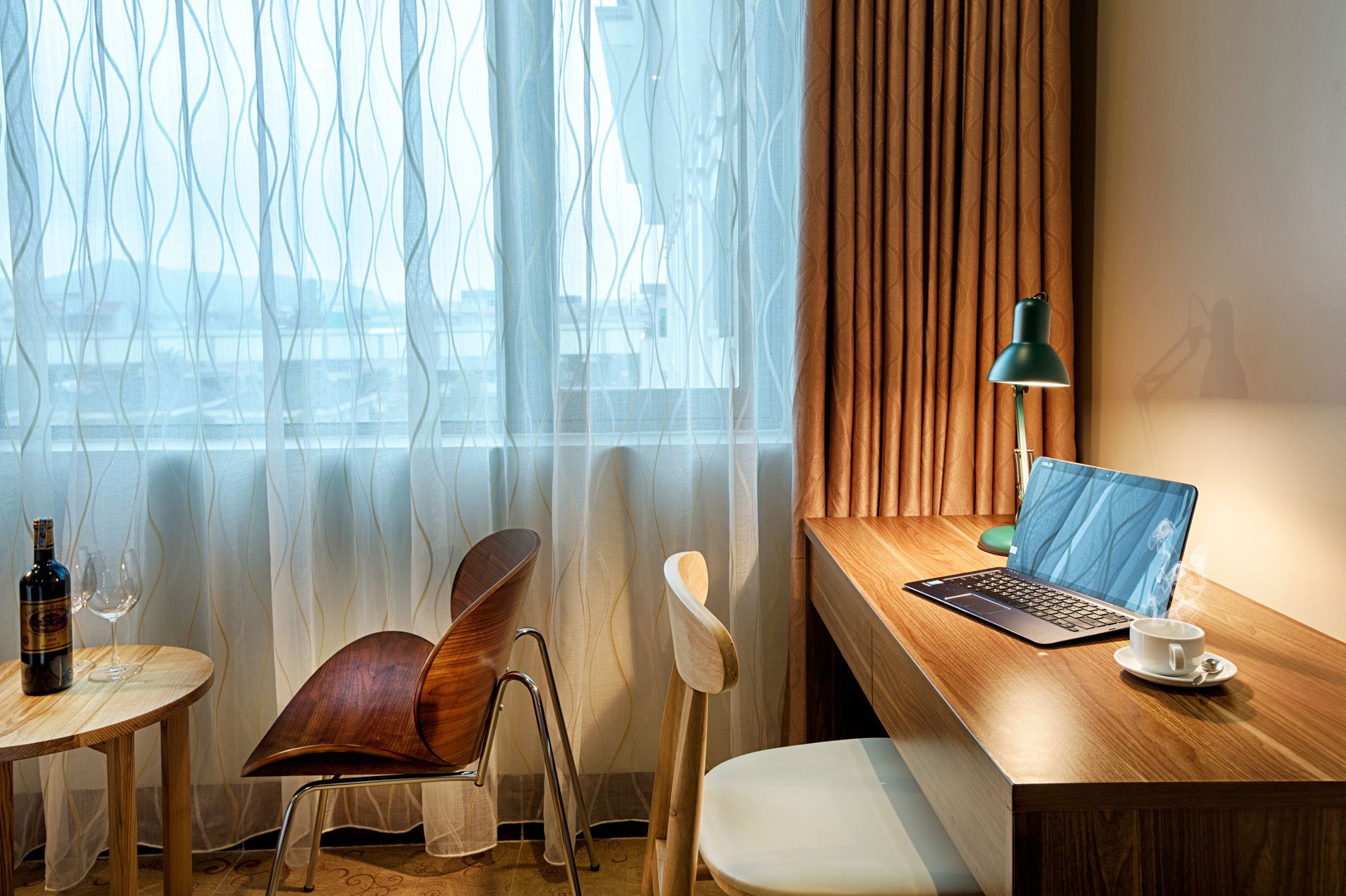 A modern hotel room with a wooden desk, a laptop, a green desk lamp, a white cup, and a bottle of wine on a small table.