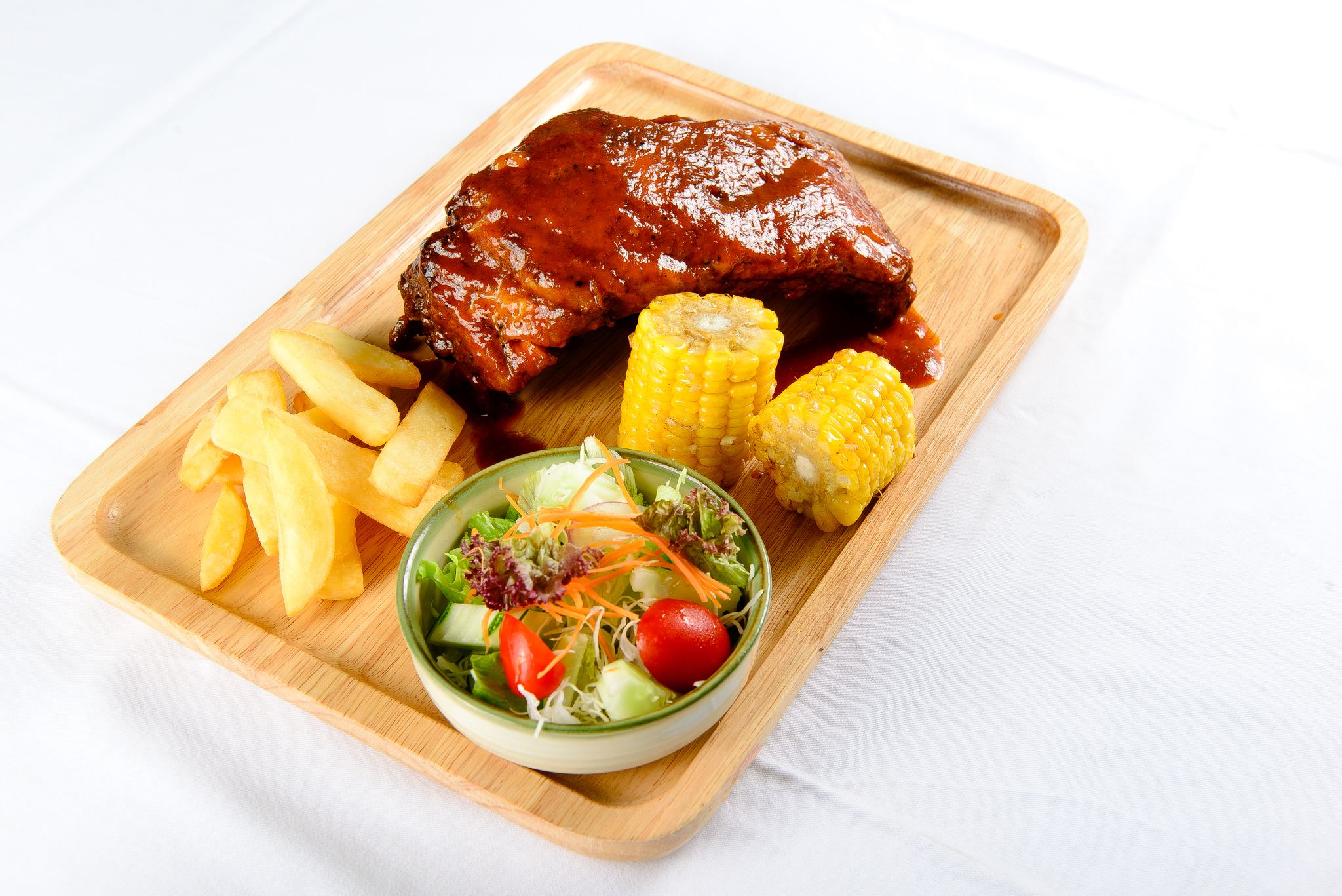 A wooden serving board with a grilled meat, French fries, corn on the cob, and a salad.