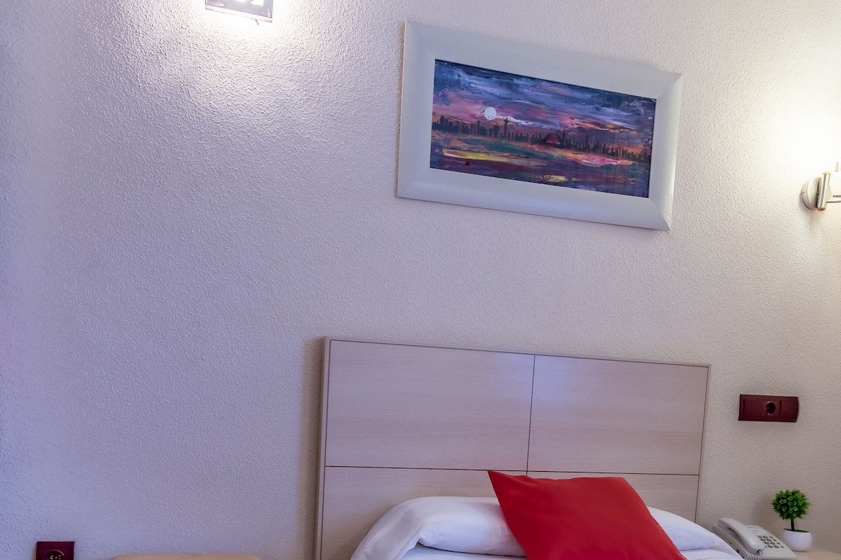 Quarto de hotel bem arrumado com uma cama, uma mesinha, uma mesa de cabeceira com telefone e um quadro na parede.