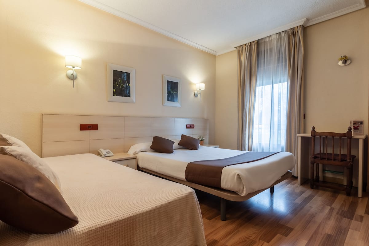 Quarto de hotel com duas camas, uma escrivaninha e uma cadeira. Há luminárias fixadas na parede e quadros emoldurados.