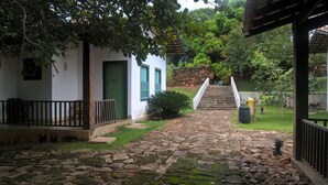 A stone pathway leading to a staircase surrounded by greenery and a white building with green doors. - Pousada do Ipê (Goiás)