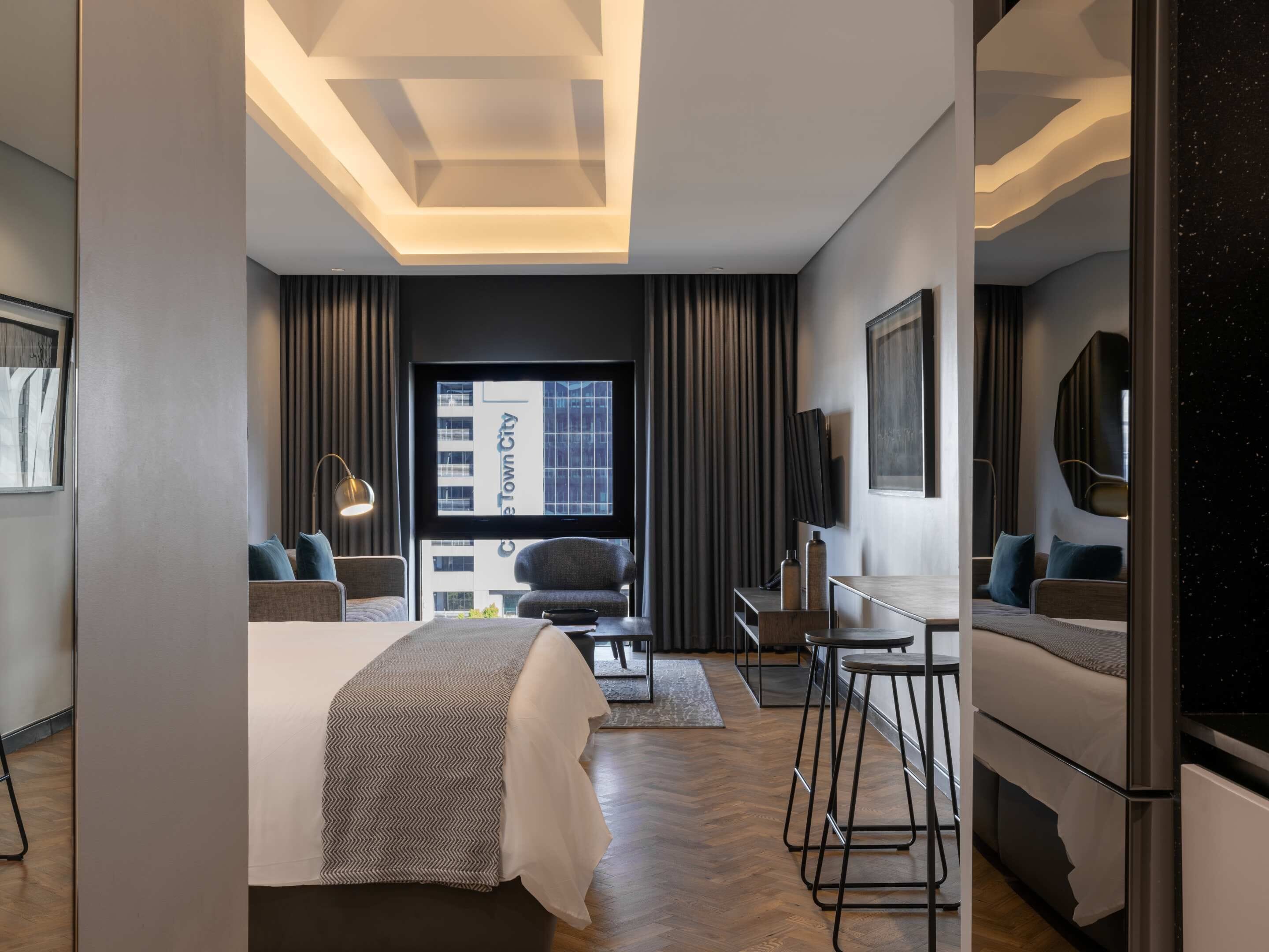 A modern hotel room with a large bed, a desk, a chair, and a mirror. There is a window with a view of a building outside.