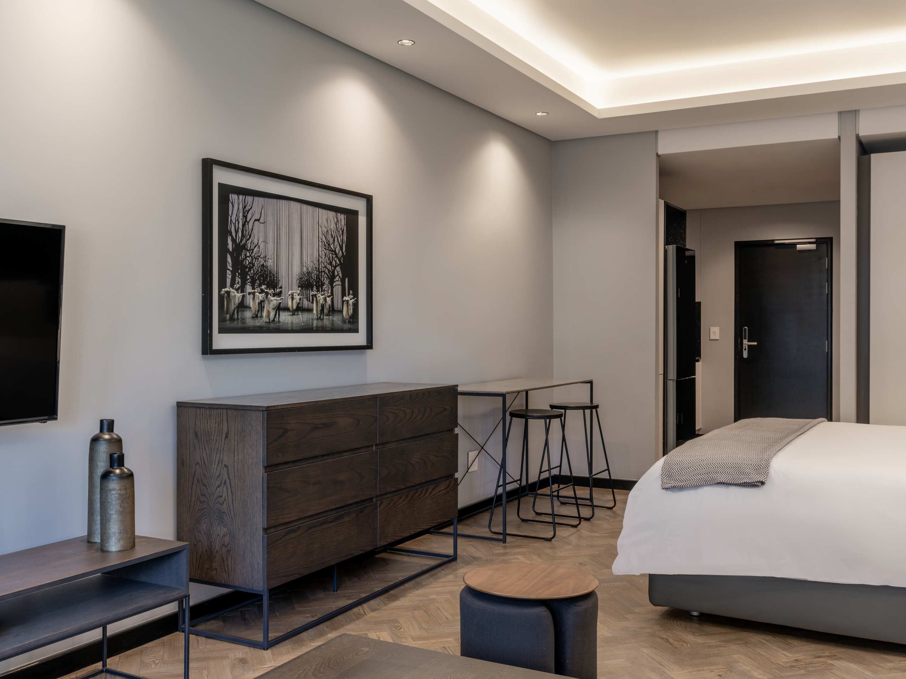 Une chambre d’hôtel moderne avec un grand lit, une commode en bois, un bureau avec deux tabourets, une œuvre d’art encadrée au mur et un téléviseur sur le côté gauche.