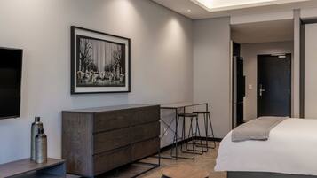 Una habitación de hotel moderna con una cama grande, un aparador de madera, un escritorio con dos taburetes, una obra de arte enmarcada en la pared y un televisor a la izquierda.