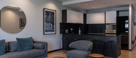 A modern living room with a grey sofa, a round coffee table, and a small round ottoman. There is a kitchen area with a black countertop and white cabinets.