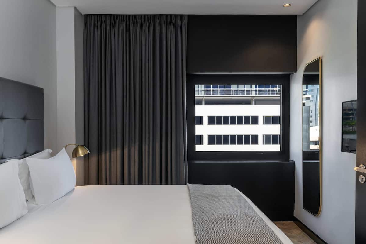 Une chambre d’hôtel moderne avec un grand lit, une fenêtre donnant sur des immeubles, un rideau foncé et un miroir fixé au mur.