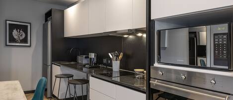 A modern kitchen with white cabinets, a black countertop, and a stainless steel microwave. There is a framed picture on the wall and a teal chair at the dining table.