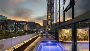 Un espace piscine sur le toit, avec un jacuzzi, des chaises longues et une vue sur la ville au crépuscule.