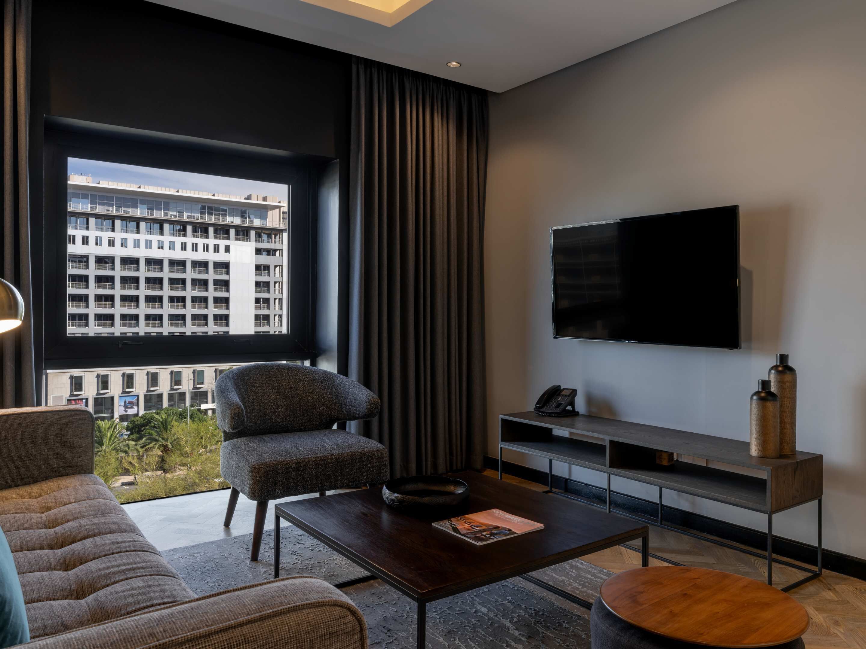 A modern living room with a large window, a flat-screen TV, a sofa, a coffee table, and a side table with a phone and decorative bottles.