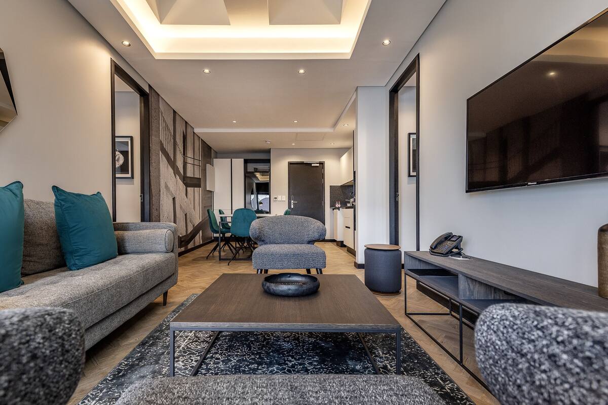 Un salon moderne avec un canapé, un pouf et une table basse. On y trouve également un coin repas avec des chaises et une table, ainsi qu’une cuisine équipée d’un évier et d’un téléphone.