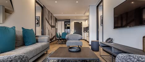 A modern living room with a sofa, ottoman, and a coffee table. There is a dining area with chairs and a table, and a kitchen area with a sink and a telephone.