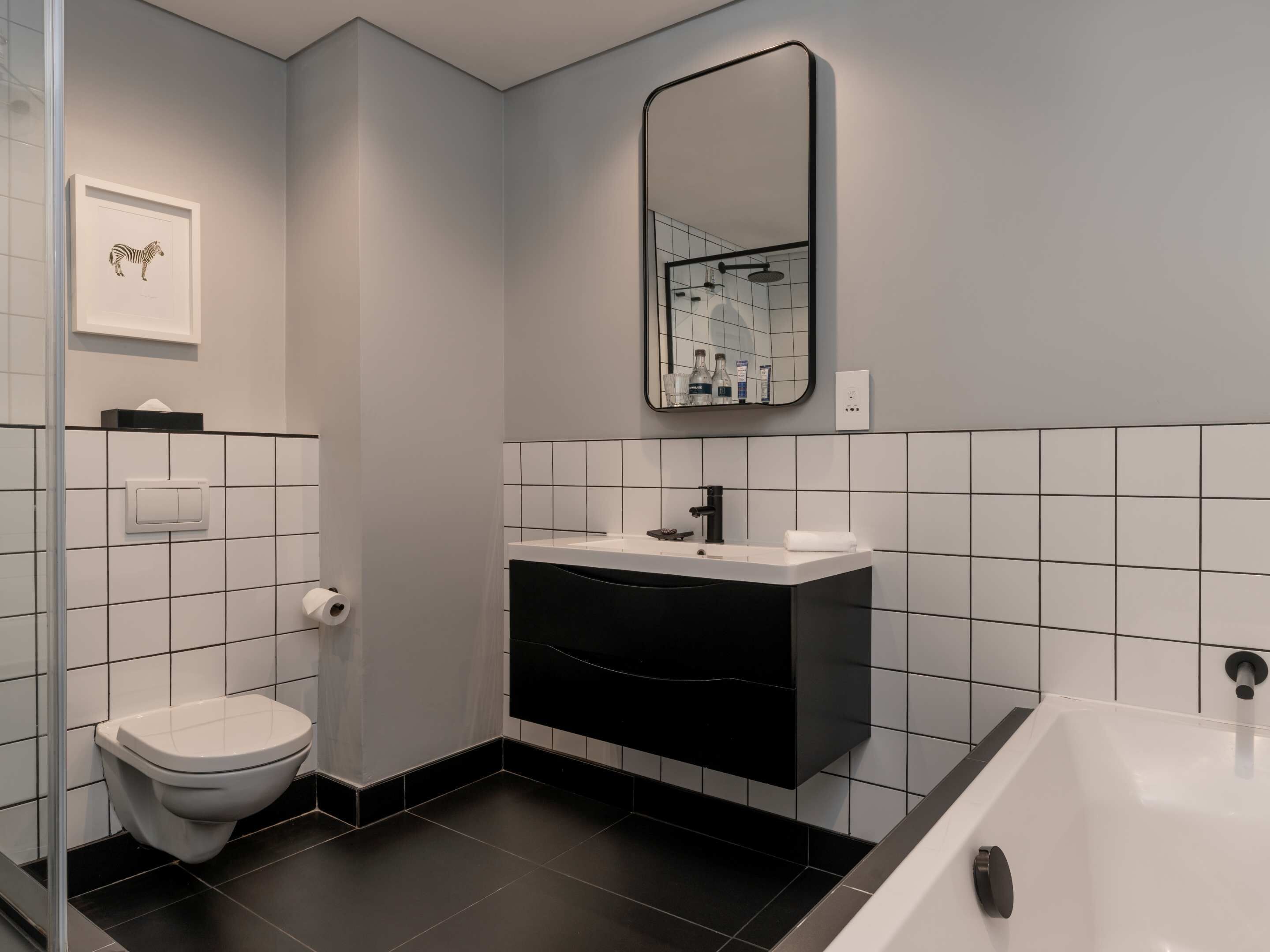 A modern bathroom with a toilet, sink, and bathtub, featuring black and white tiles and a framed zebra artwork on the wall.