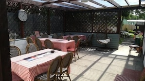 Une terrasse extérieure avec des tables et des chaises, protégée par un toit transparent, et une horloge au mur.