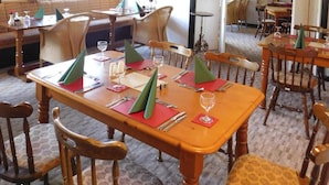 Un espace repas avec des tables et des chaises en bois, un plafond haut avec des poutres apparentes et une fenêtre avec des rideaux.