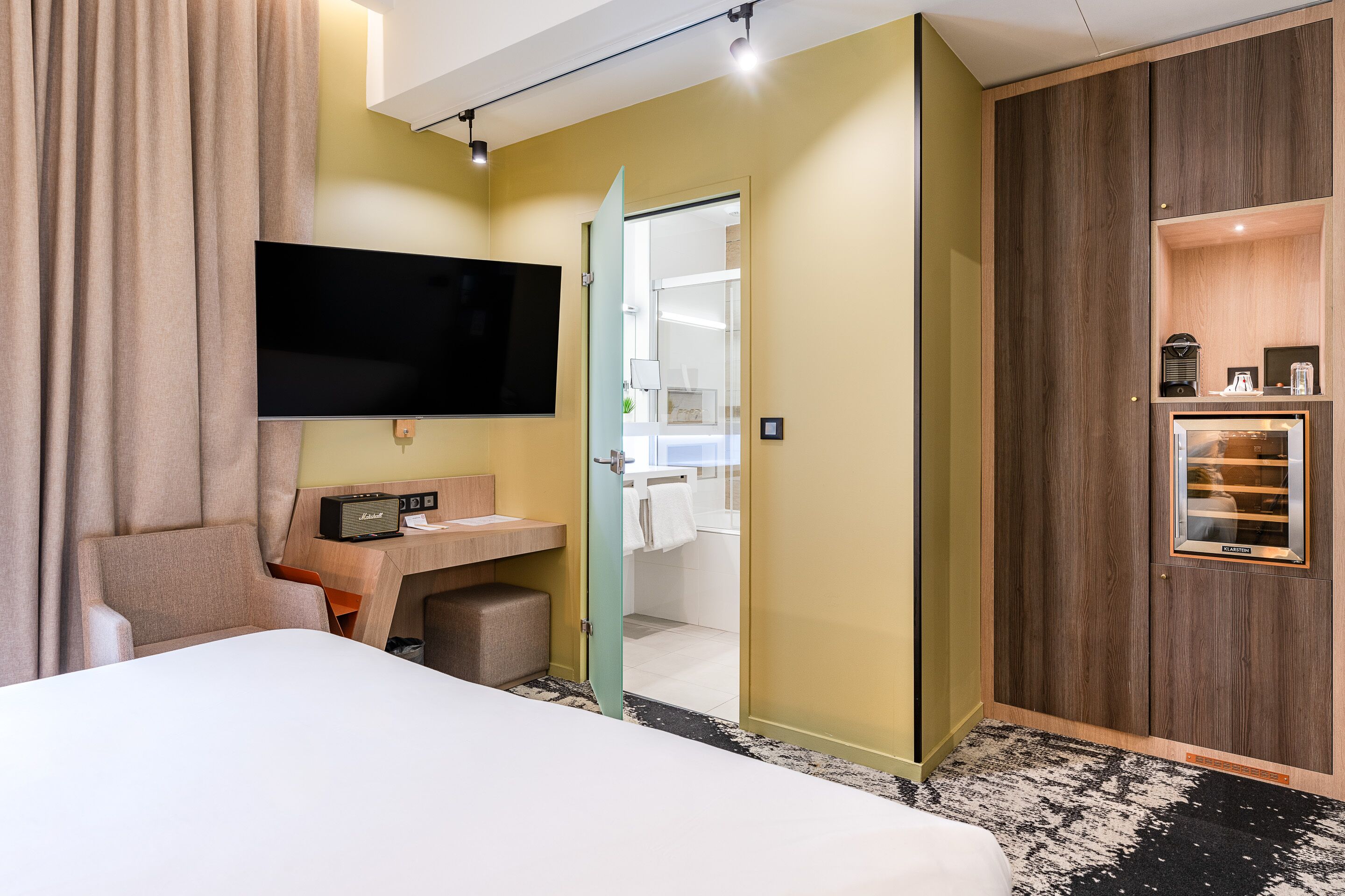 Una camera d'albergo con un letto, una scrivania, una TV a schermo piatto, un minibar e un bagno visibile attraverso una porta aperta.