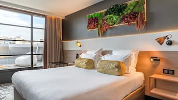 A modern hotel room with a large bed, a view of the cityscape through floor-to-ceiling windows, a decorative wall with greenery, and a small bedside table with a lamp.