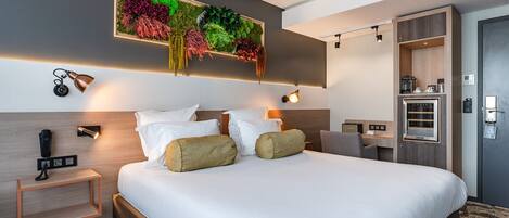 Una habitación de hotel moderna con una cama grande, una planta en la pared, lámparas de noche, una mesita pequeña y un armario empotrado.