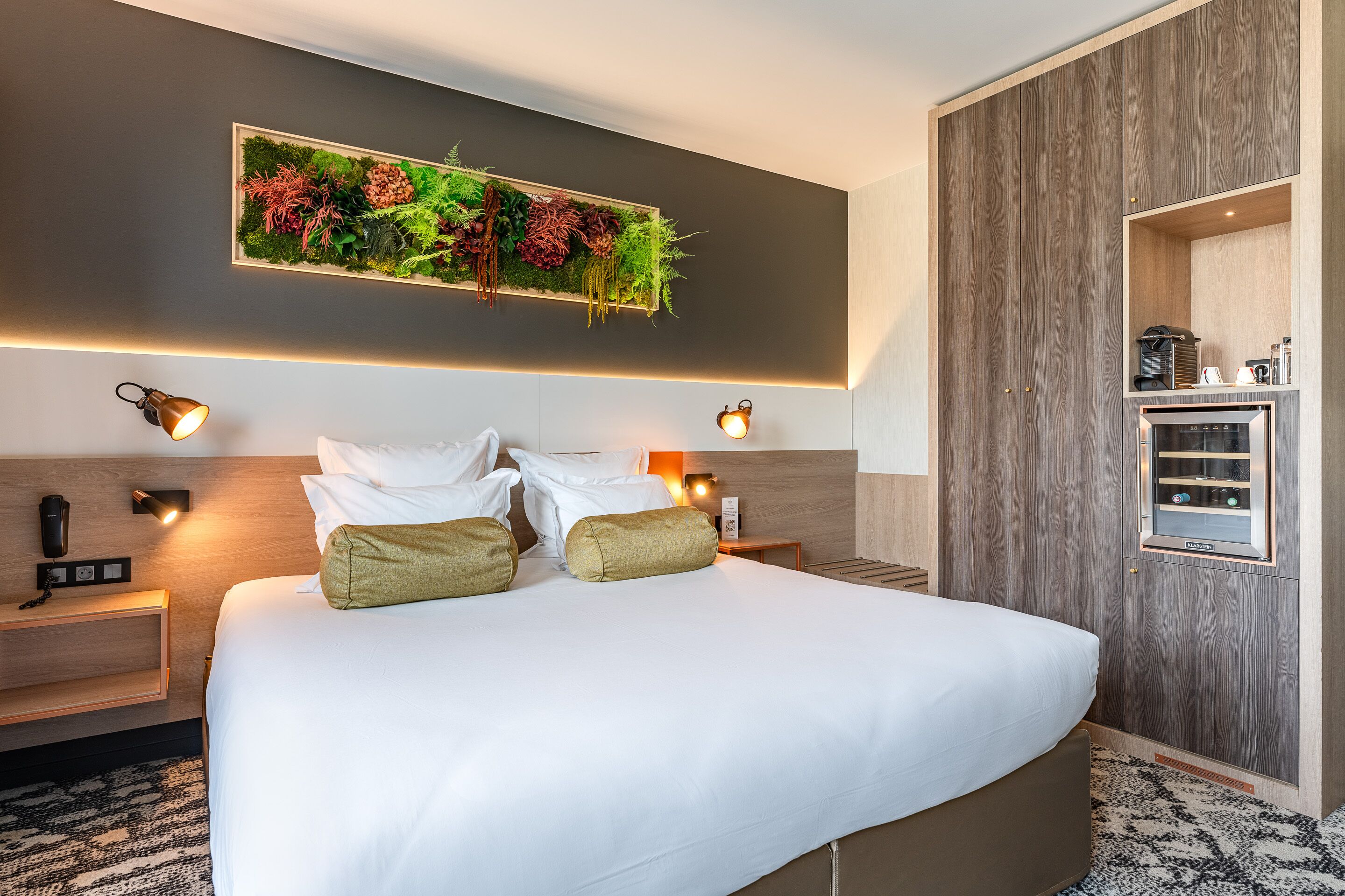 A modern hotel room with a large bed, two bedside tables, a built-in shelf with a coffee machine, and a wall-mounted plant.