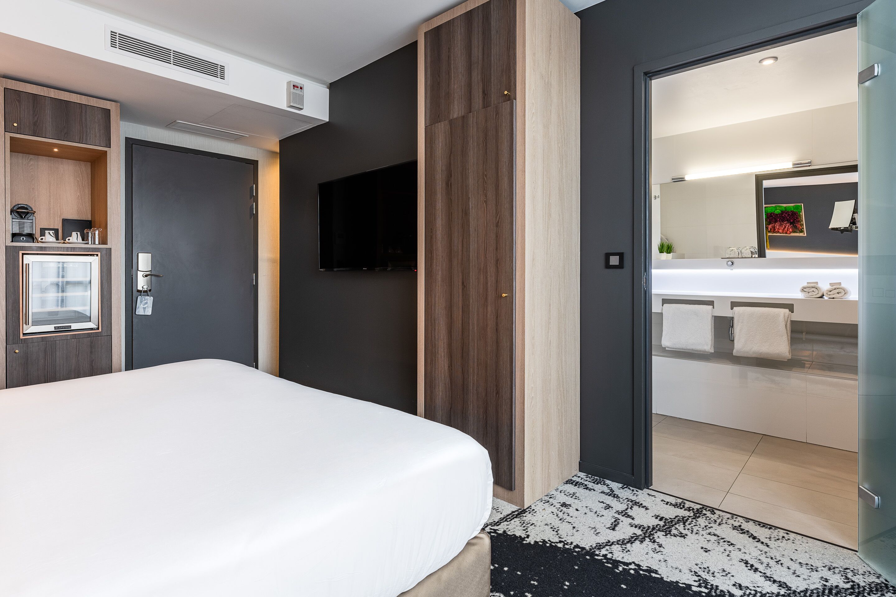 A modern hotel room with a large bed, a television, a built-in wardrobe, and a bathroom with a large mirror and sink.