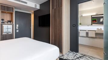 Una habitación de hotel moderna con una cama grande, televisión, armario empotrado y baño con un espejo grande y lavabo.