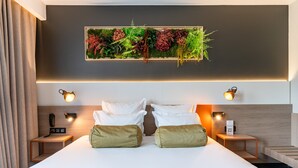 Ein modernes Hotelzimmer mit einem großen Bett, zwei Nachttischen, einer Wandpflanze und einem Telefon auf der linken Seite.