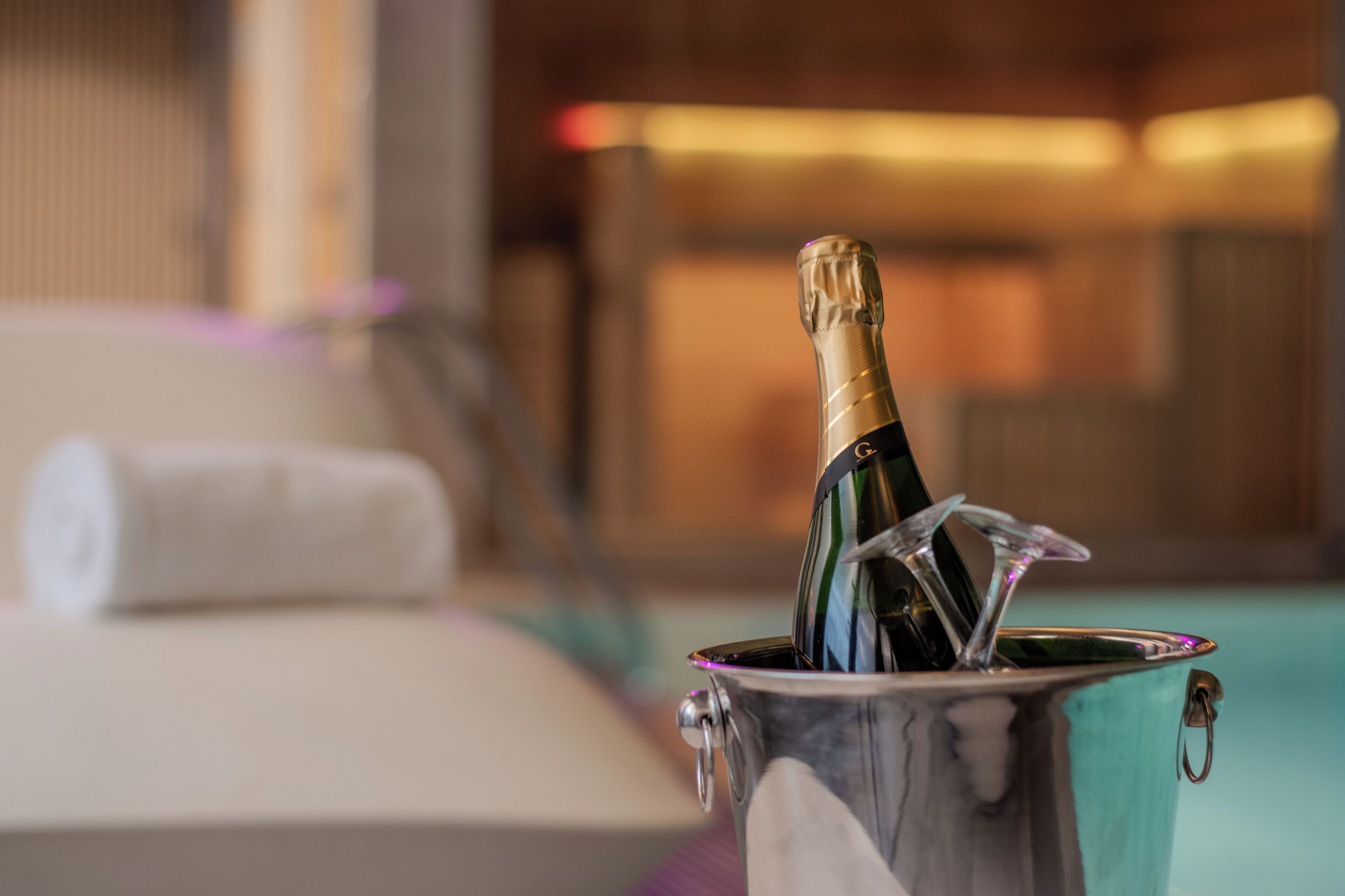 A bottle of champagne in a metallic bucket, with a towel and a blurred background suggesting a hotel setting.
