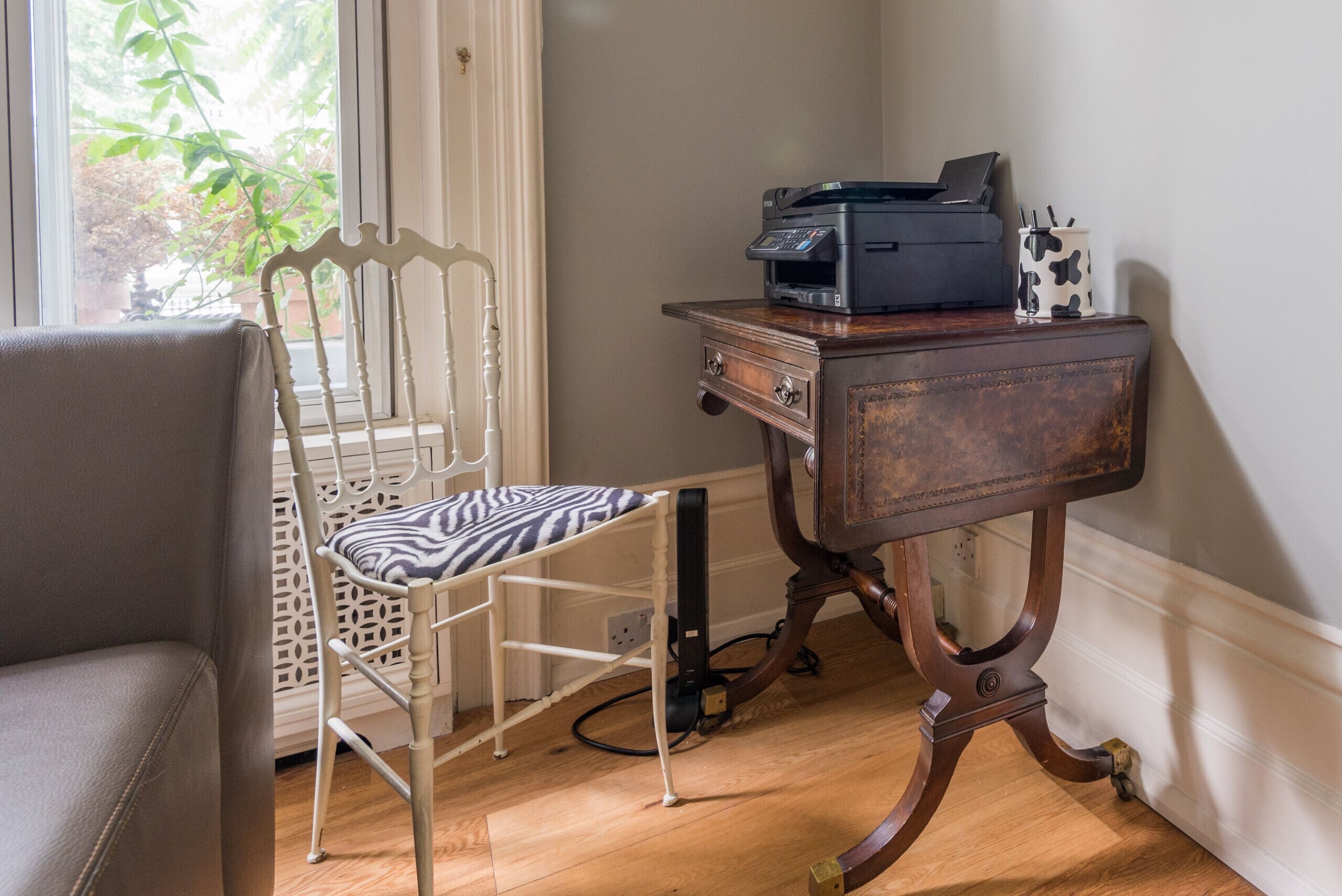 A room with a white chair with a zebra-patterned cushion, a wooden side table with a printer, and a window with a view of greenery.