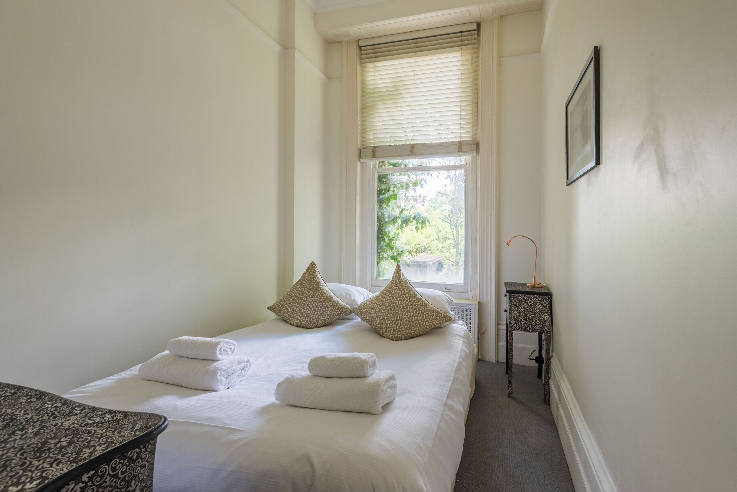 A neatly made bed with two pillows, a bedside table, and a window with blinds.