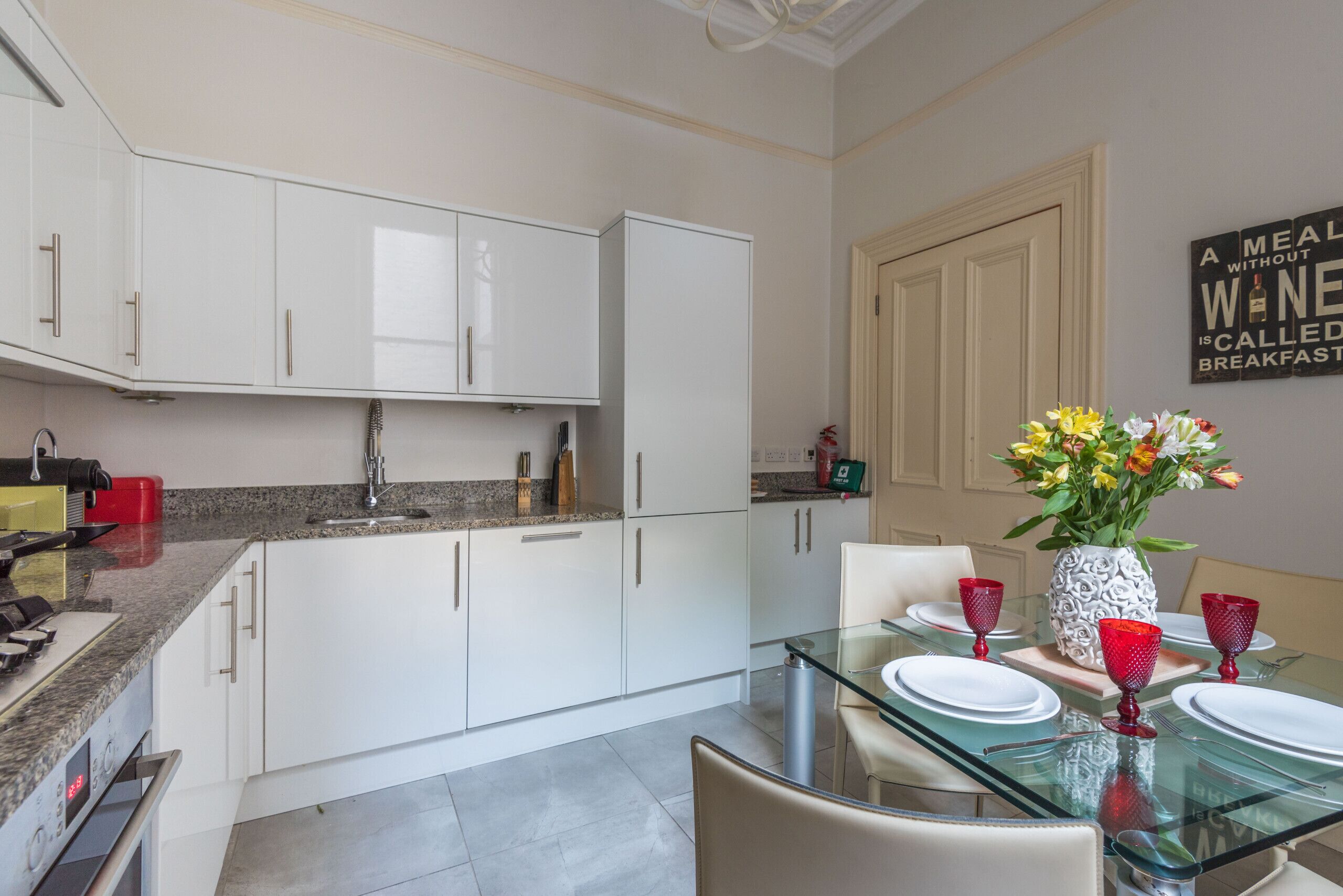 Una cocina moderna con armarios blancos, encimera de granito y comedor con mesa de vidrio y cristalería roja.