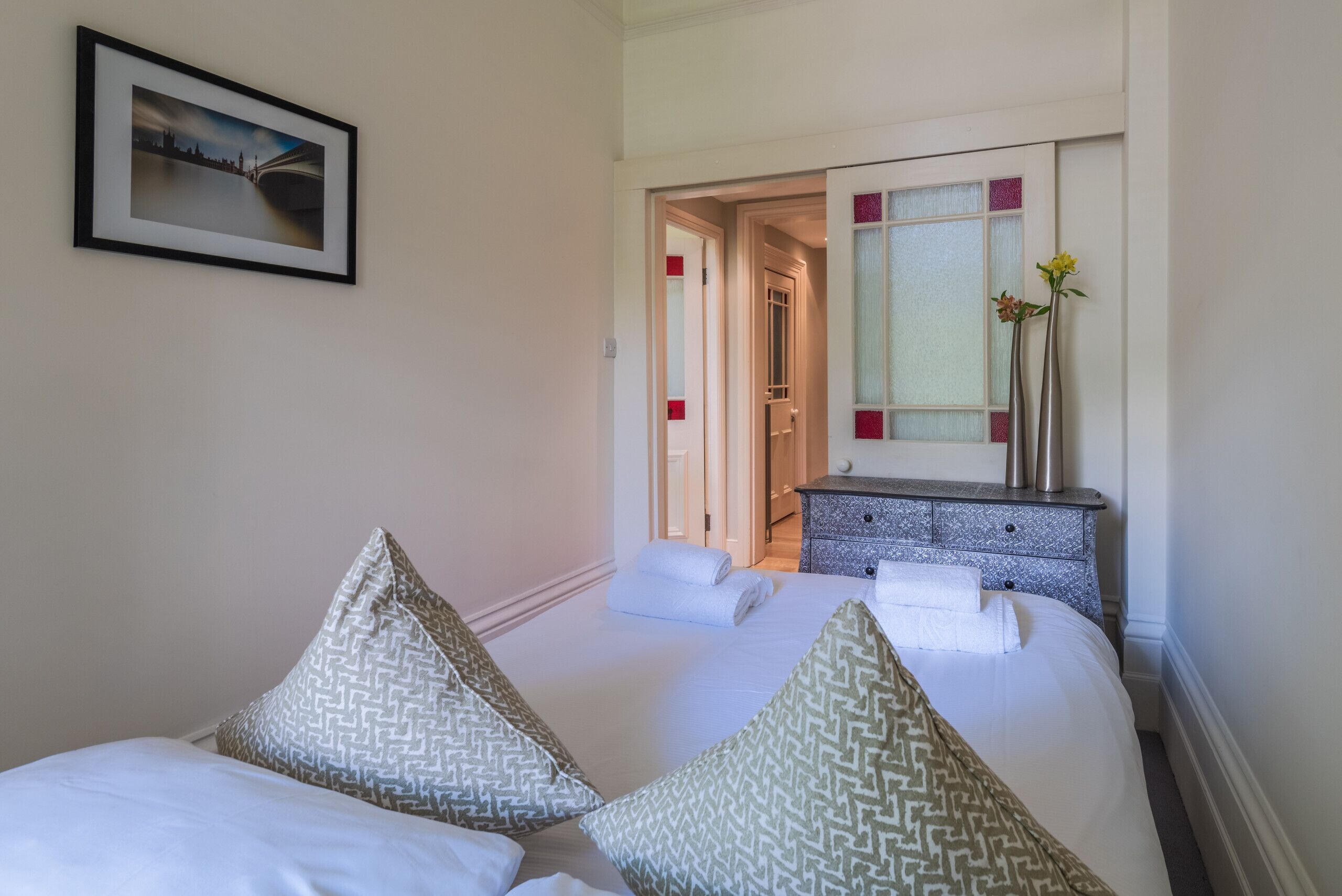 Habitación de hotel con cama, almohadas, mesita de noche, espejo y un cuadro en la pared.