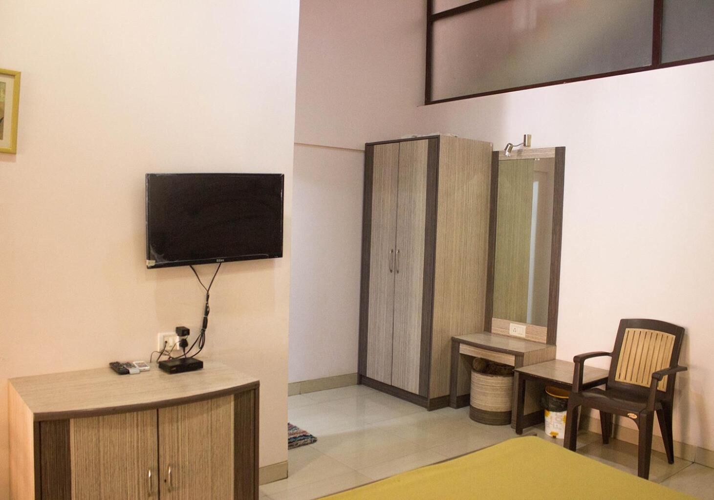 A bedroom with a TV mounted on the wall, a wardrobe, a bedside table, a chair, and a mirror.