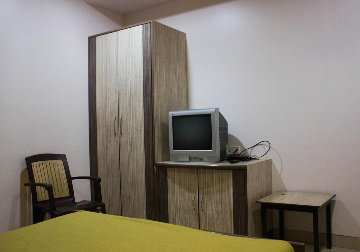 A bedroom with a bed, a wooden wardrobe, a television on a wooden stand, a chair, and a small table.