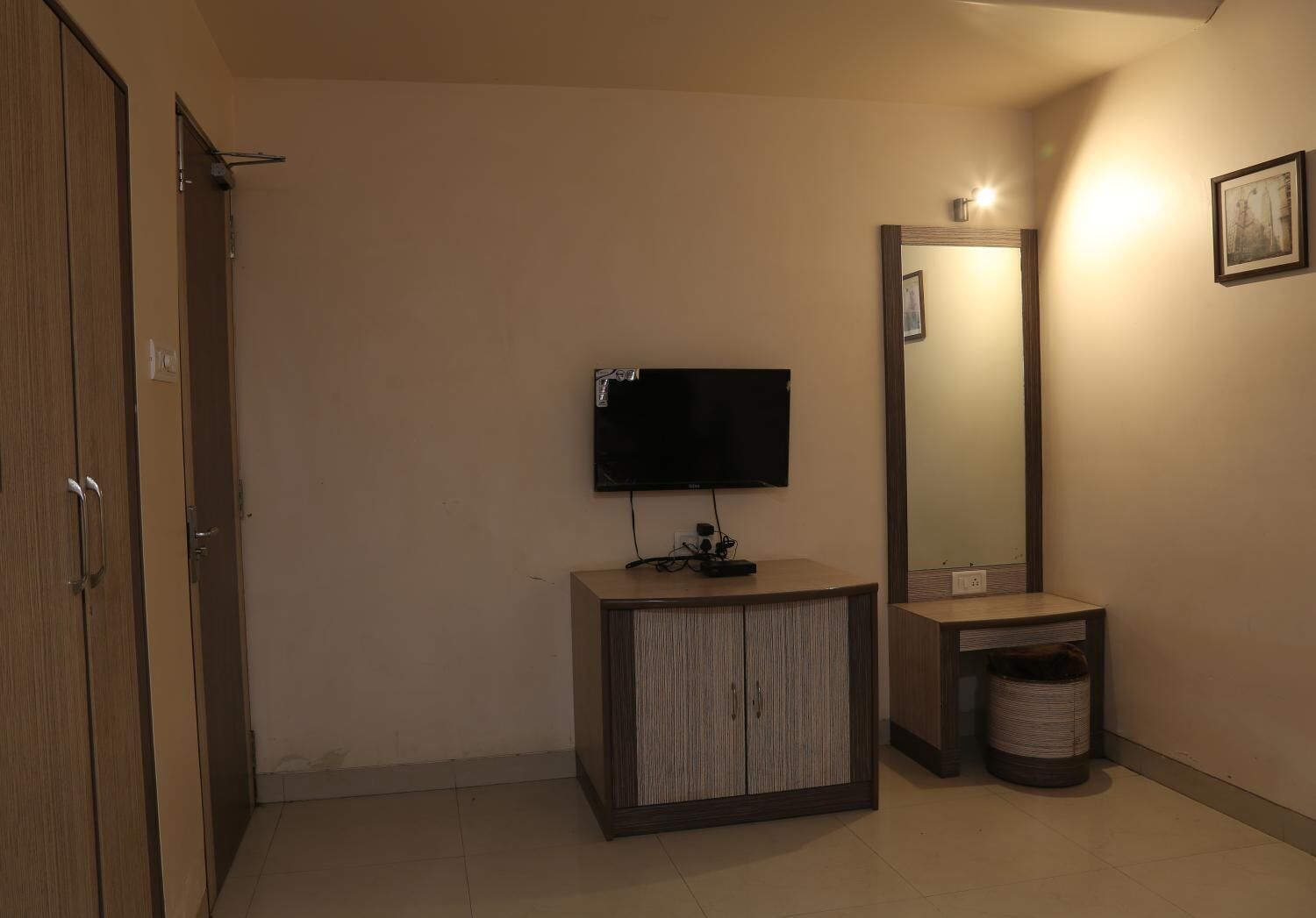 Quarto de hotel com guarda-roupa de madeira, um pequeno armário de madeira com uma televisão embutida acima, um espelho e uma lixeira.